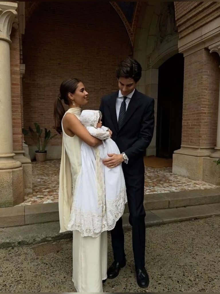 Elegant couple holding a baby at a traditional event in front of a historic brick building.