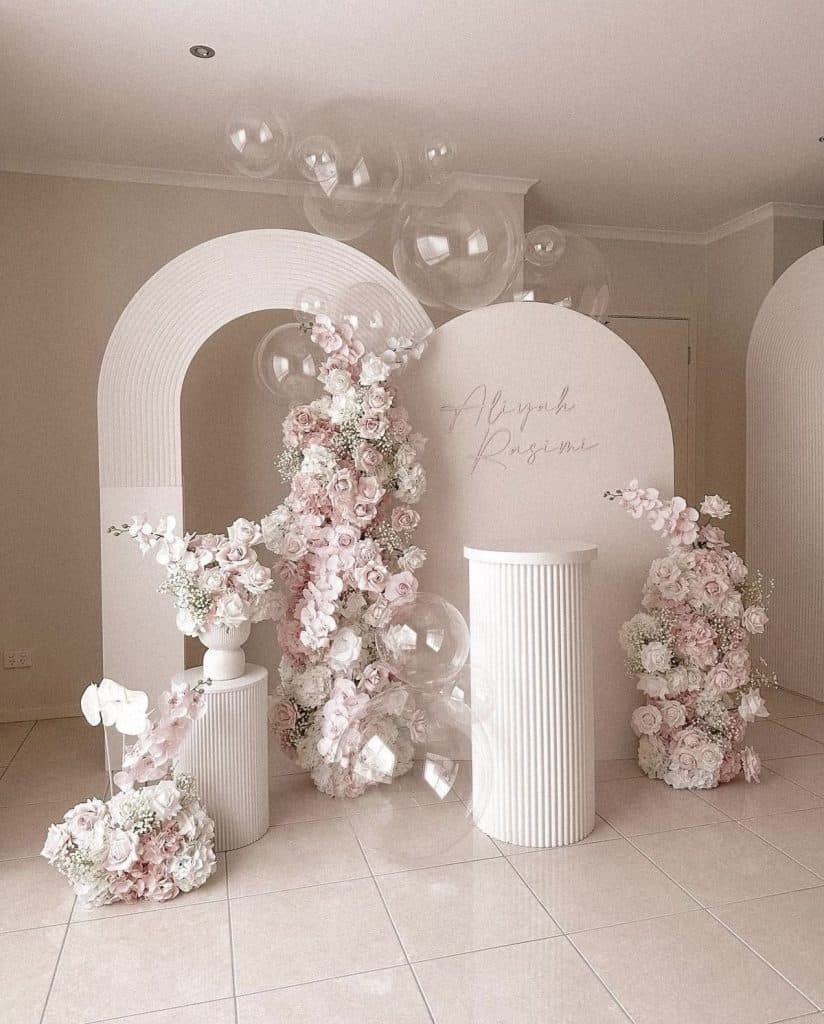 Elegant wedding backdrop with pink and white floral arrangements, balloons, and personalized signage.