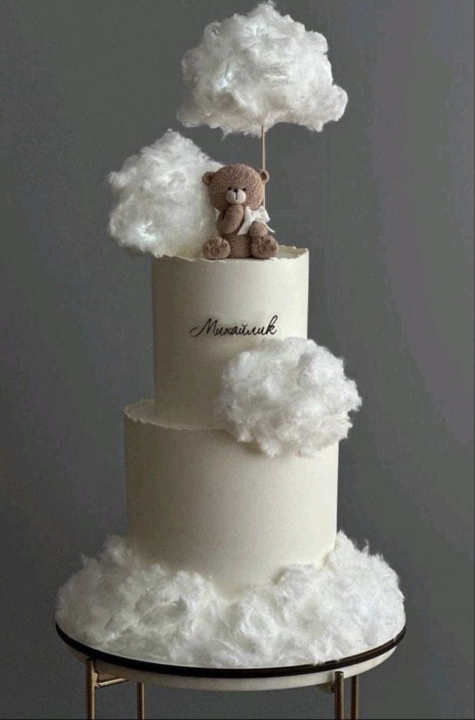 Fluffy cloud-themed birthday cake with teddy bear topper, elegant white icing, and personalized inscription.
