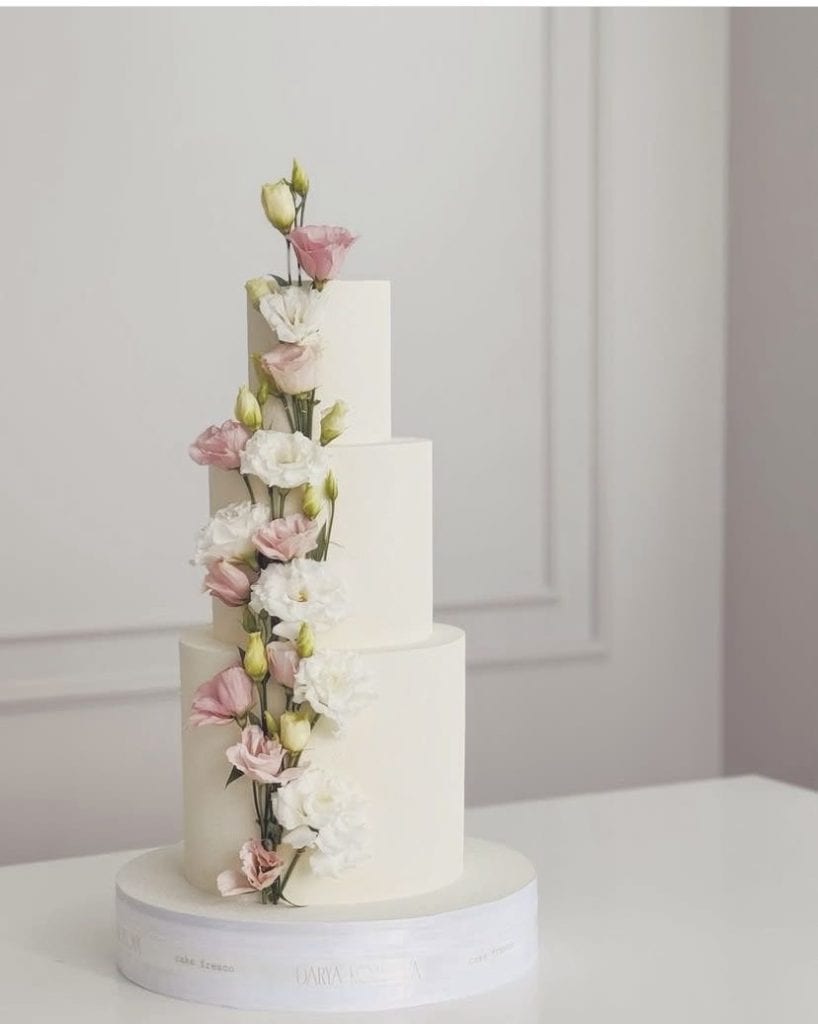 Elegant three-tier white wedding cake decorated with pink and white lisianthus flowers for special events and celebrations.