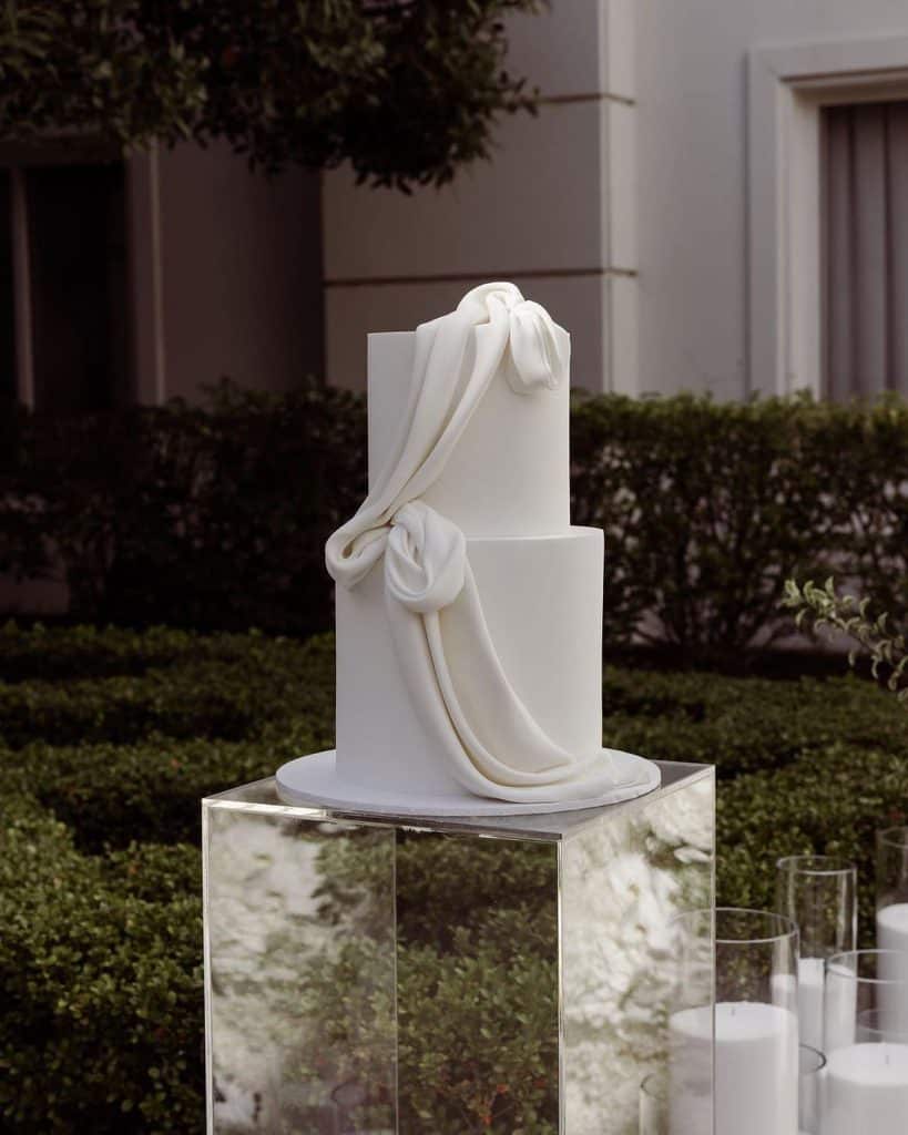 Elegant wedding cake with white fondant and draped fondant ribbons, displayed outdoors.