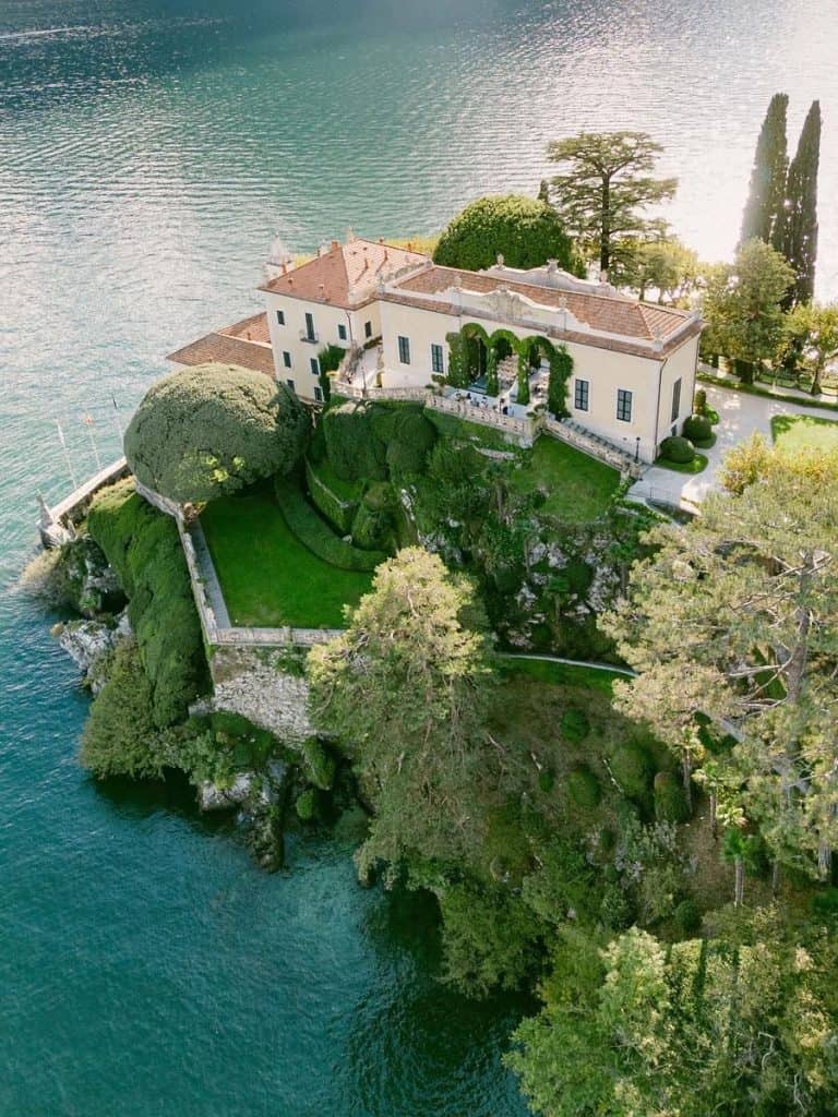 Stunning aerial view of a lakeside mansion surrounded by lush greenery and overlooking calm water.