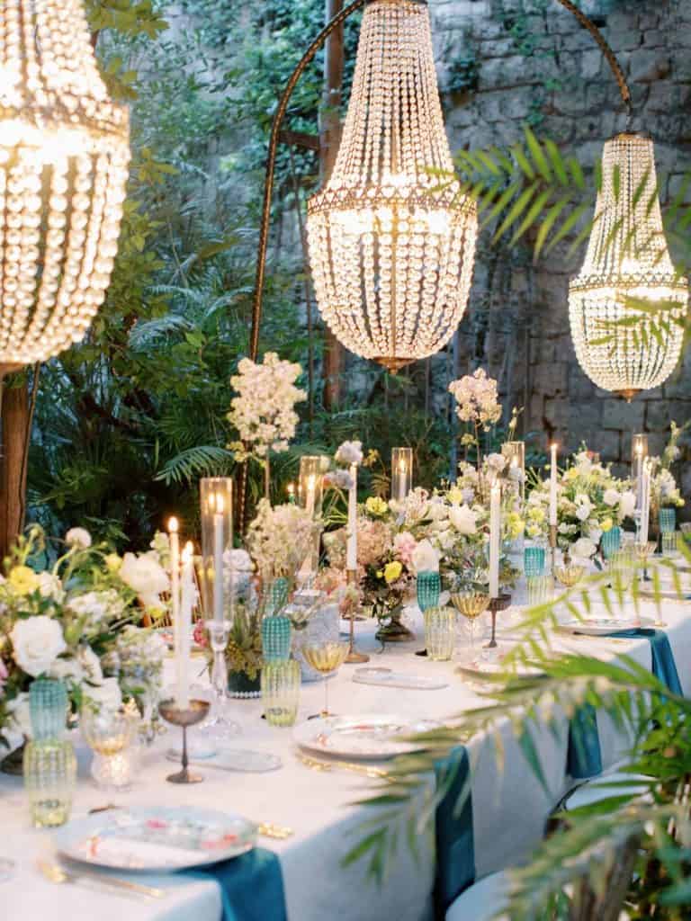 Elegant outdoor event table with floral centerpieces and chandelier lighting, perfect for luxury weddings or garden parties.