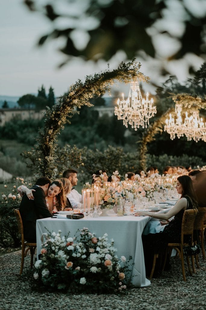 Elegant outdoor wedding reception under chandeliers with floral decor and candlelit table setup.