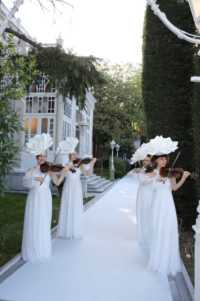 Elegant women in white dresses with flower hats playing violins at outdoor wedding or event.
