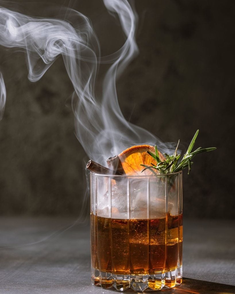 Warm amber cocktail garnished with orange slice and rosemary, with swirling steam, on a dark background.