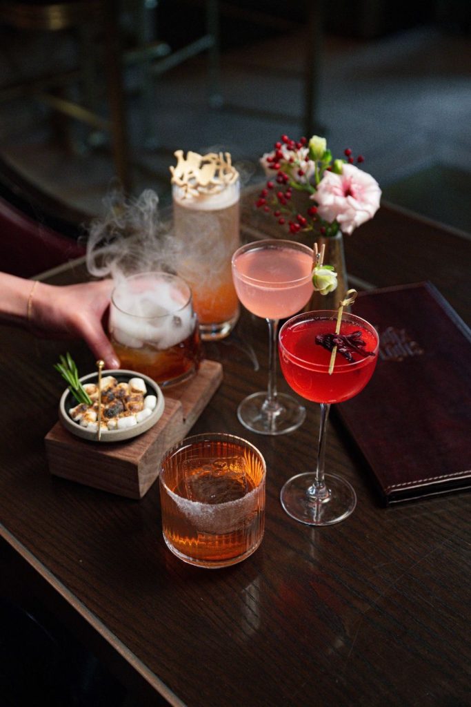 Vibrant cocktail drinks on a dark wooden table with flowers and snacks, perfect for event or celebration atmospheres.