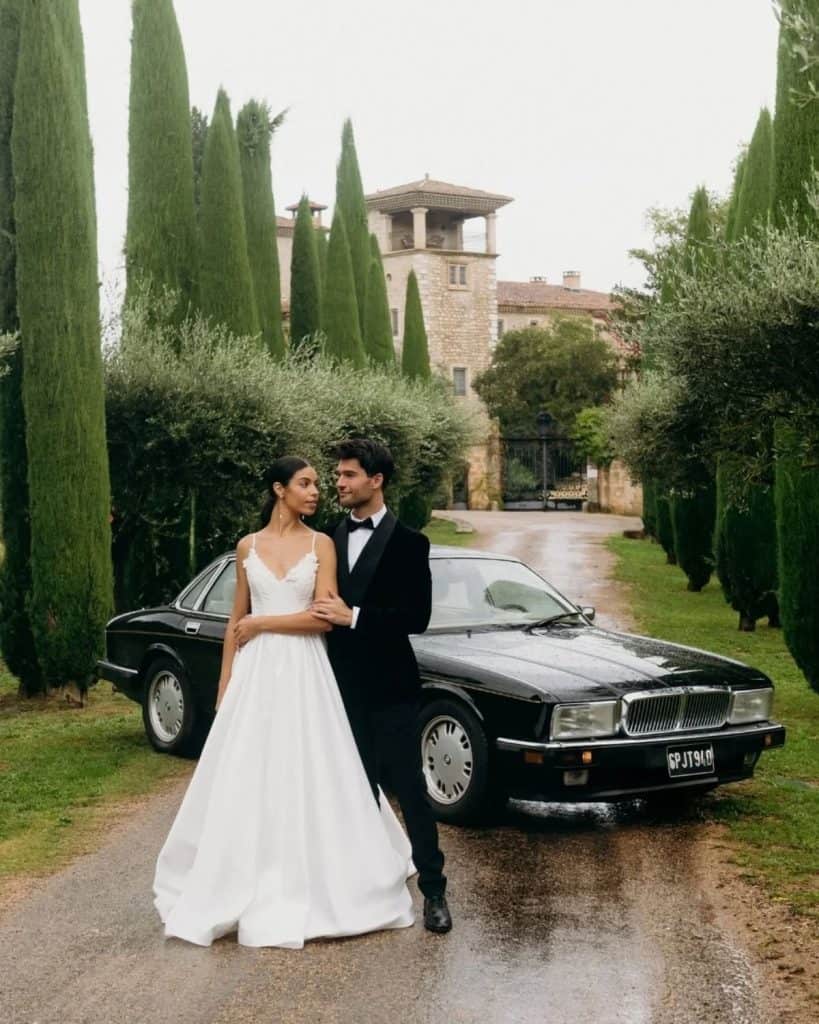 Elegant wedding couple posing beside a luxury classic car in a picturesque estate setting.