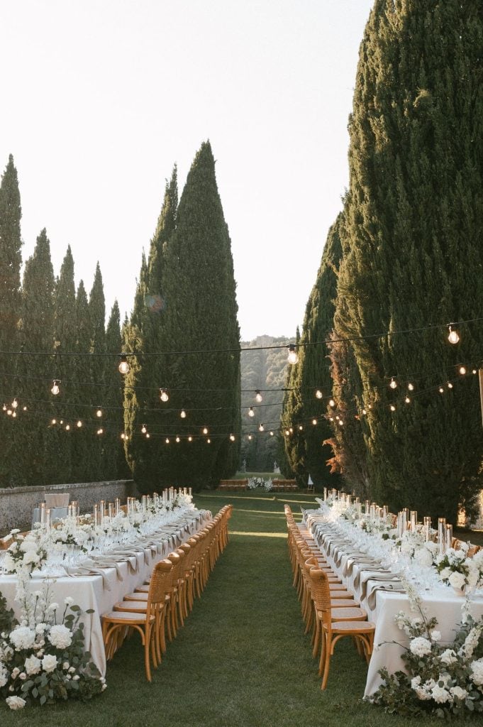 Elegant outdoor wedding reception setup with long banquet tables, floral decor, and string lights amid tall cypress trees.
